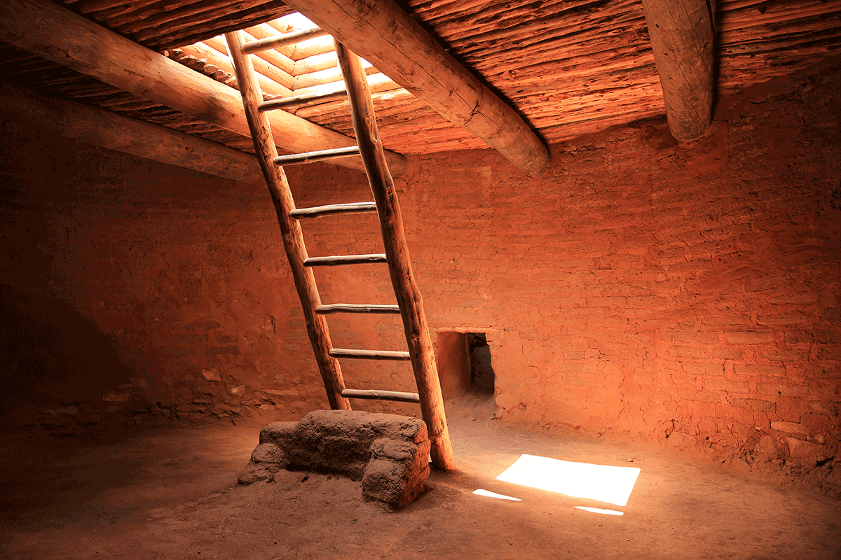 A wooden ladder leaning into an opening of the ceiling in a Native American kiva with sunlight pouring in.